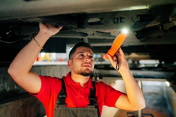 Ein Azubi ist in der Werkstatt unter einem Bus und leuchtet mit einer Taschenlampe. 