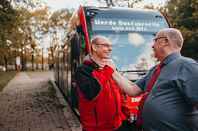 Zwei Busfahrer klatschen zusammen ein, im Hintergrund steht ein Bus mit der Zielanzeige Werde Busfahrer*in.