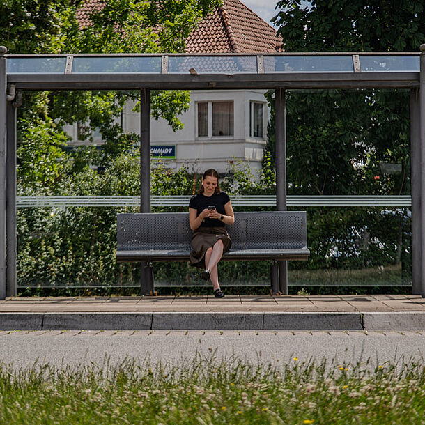 Frau sitzt an einer Bushaltestelle mit einem Handy in der Hand