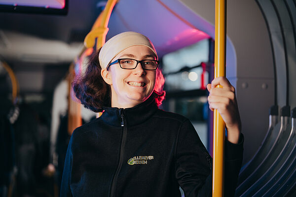 Berufskraftfahrerin steht im Innenraum eines Busses, hält sich an einer Haltestange fest und lächelt.