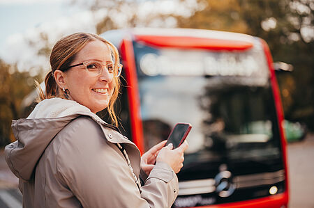 Frau hält ihre digitale Fahrkarte in der Hand und steht vor dem Bus.
