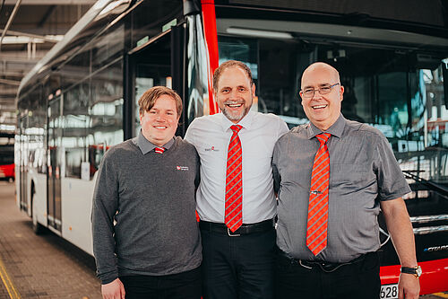 Drei Mitarbeitende stehen vor einem Linienbus in der Werkstatthallte und halten sich in den Armen.