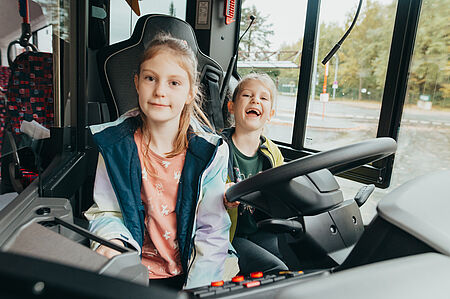 Zwei Kinder sitzen im Bus hinterm Steuer und haben Spaß und lachen in die Kamera.