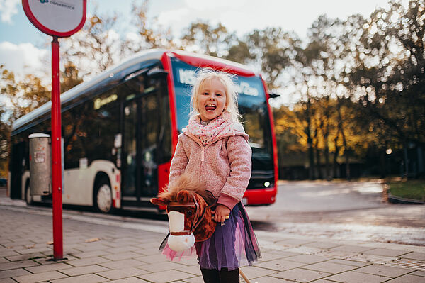 Ein verkleidetes Kind steht mit ihrem Steckenpferd an der Bushaltestelle und lacht, im Hintergrund ist ein Bus.
