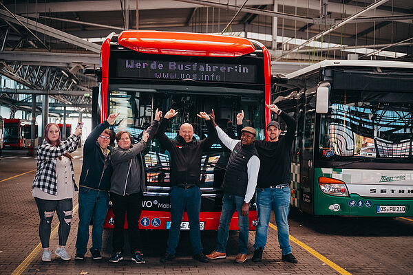 Viele Männer und Frauen stehen vor dem Bus und zeigen auf die Zielanzeige mit Werde Busfahrer*in.