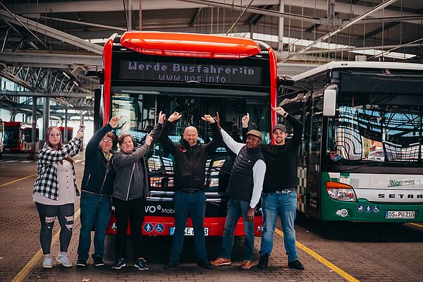 Viele Busfahrer und Busfahrerin stehen vor einem Bus und zeigen auf die Zielanzeige mit dem Inhalt Werde Busfahrer*in bei der VOS.