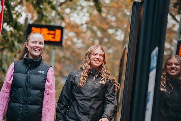 Zwei Mädchen lächeln und steigen in einen Bus ein, im Hintergrund ist eine digitale Fahrgastanzeige zu sehen.