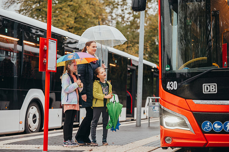 Mutter mit ihren zwei Kinder steht an der Haltestelle und möchte gleich in den Bus einsteigen.