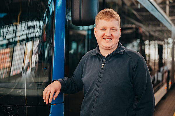 Junger Mann steht vor einem Bus, lehnt sich leicht an die Tür des Busses und lächelt in die Kamera