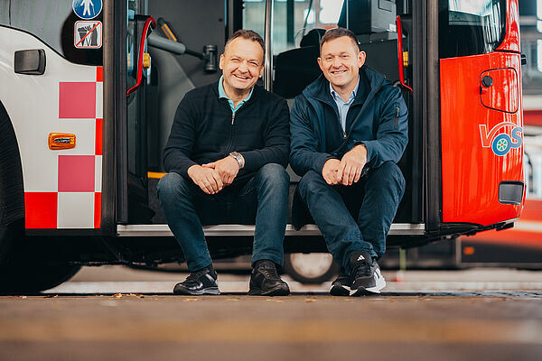 Zwei Busfahrer sitzen nebeneinander vor dem Eingang des Busses und lächeln.