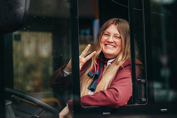 Junge Frau sitzt lächelnd am Steuer eines Busses, trägt Dienstkleidung von D B, schaut aus dem Fenster und macht ein Peace-Zeichen.