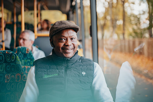 Busfahrer mit einer Mütze sitzt im Bus und lächelt, im Hintergrund Fahrgäste