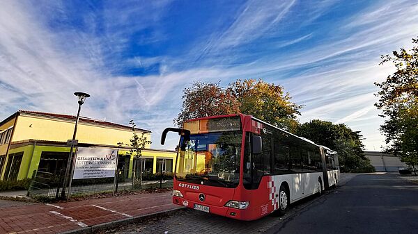 Roter Bus vor einem Schulgebäude unter blauem Himmel mit Schleierwolken.