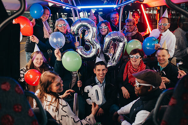 Gruppe von Mitarbeitenden und Fahrgästen feiert das 30 jährige Jubiläum der Verkehrsgemeinschaft Osnabrück im Innenraum eines Busses mit Luftballons und Jubiläumsdekoration.