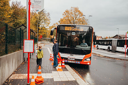 Die Bustrainerin erklärt den Kindern den Umgang an der Bushaltestelle, während der Bus sehr weit auf dem Bordstein ragt.