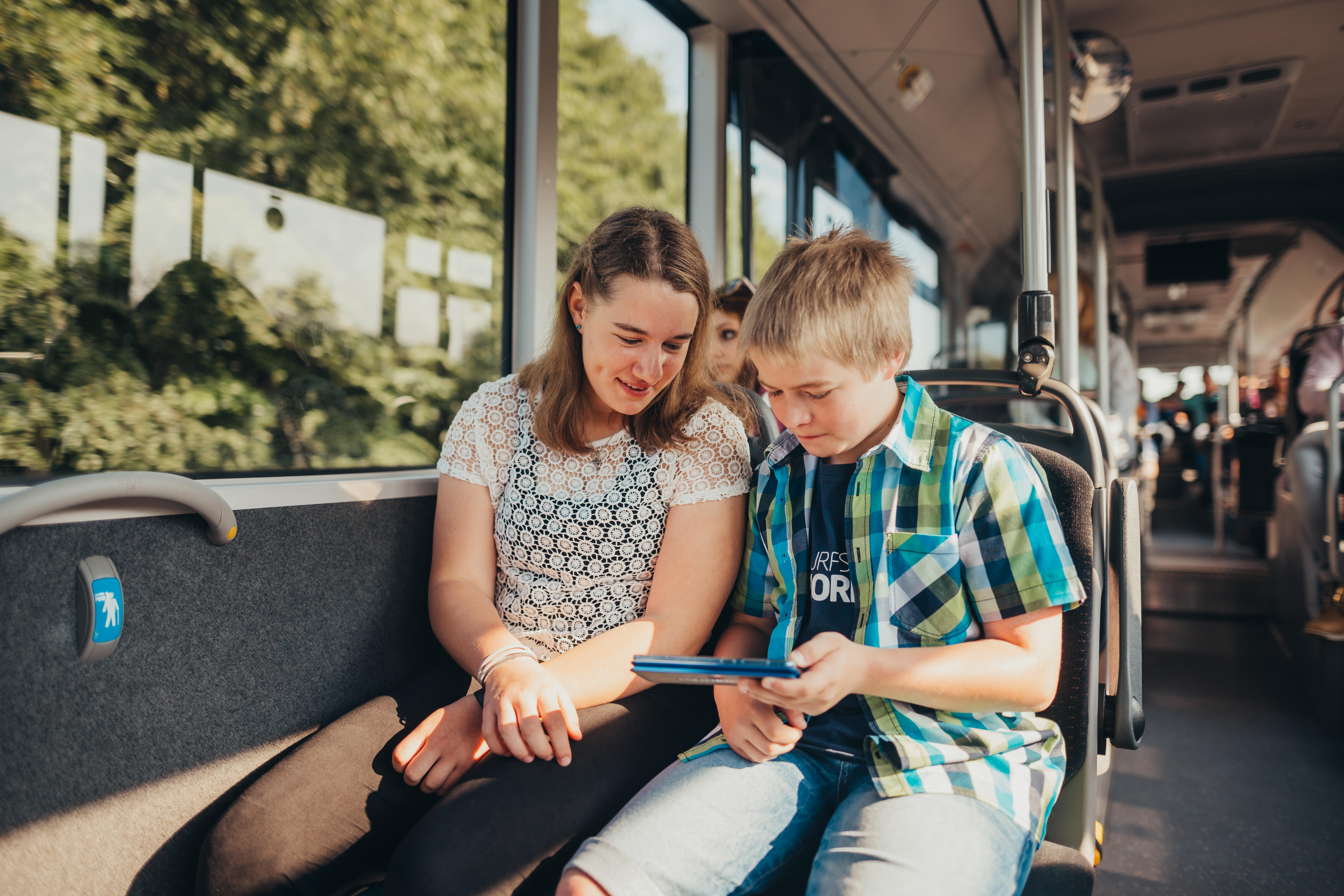 Ein Mädchen und ein Junge sitzen im Bus nebeneinander am Fenster. Der Junge hält ein Tablet in den Händen, beide schauen gemeinsam darauf und lächeln